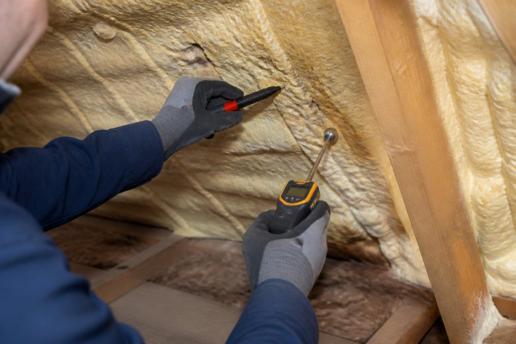 Professional surveyor's hands examining spray foam insulation in a loft space with inspection tools, pointing to areas of discoloration and checking moisture levels with a handheld meter