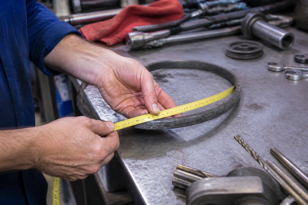 Close-up of a technician's hands wrapping a flexible tape measure around a worn V-belt removed from industrial machinery, measuring its inner circumference on a metal workshop workbench under bright fluorescent lighting