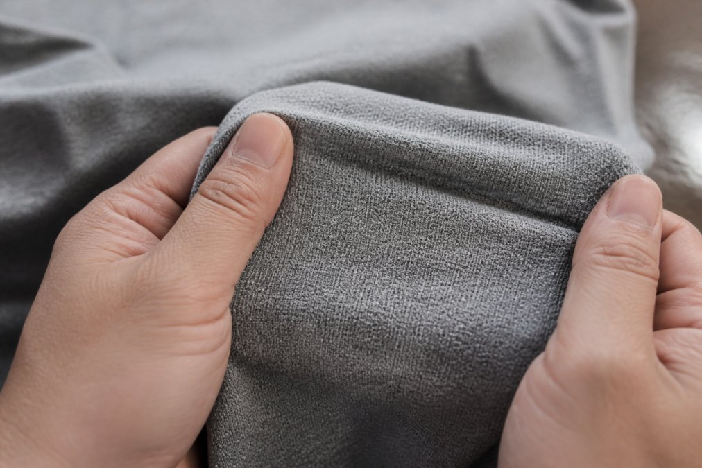Close-up of hands stretching the fabric of a heavyweight grey t-shirt between fingers, showing its dense texture and thickness under natural light