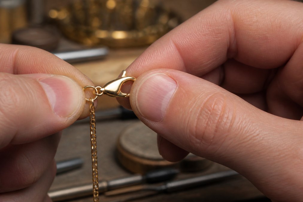 Close-up macro photograph of fingers testing the spring tension on a gold lobster claw clasp, jewelry workbench softly blurred in the background