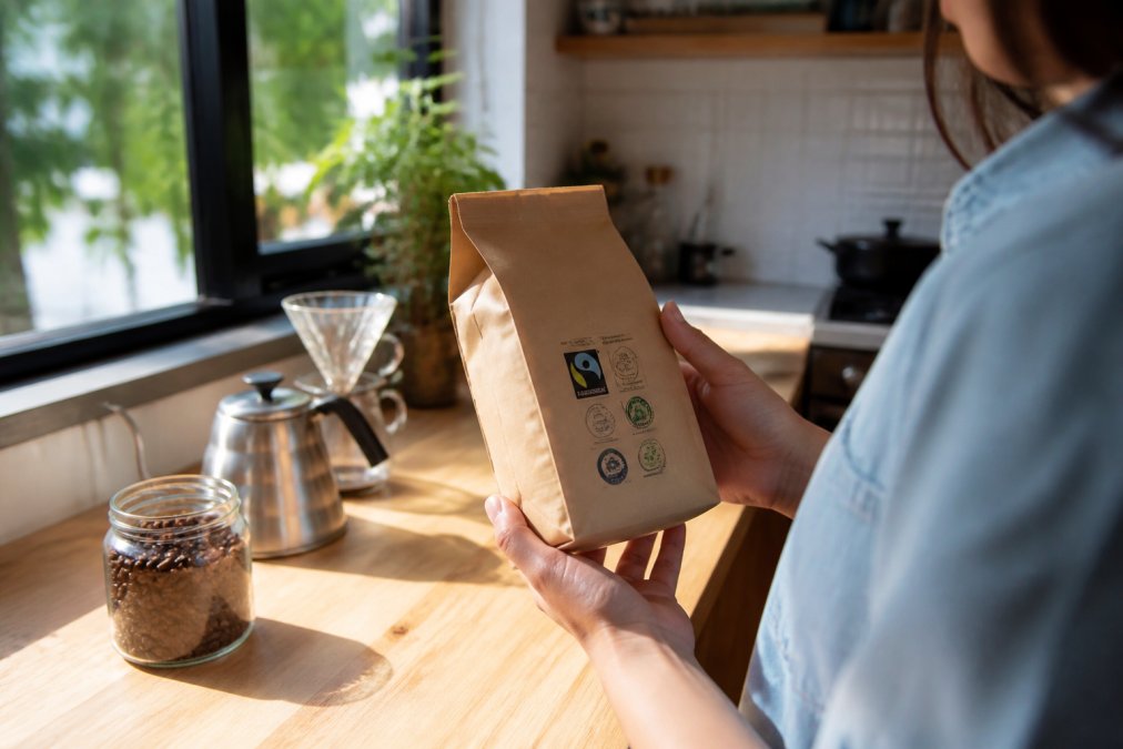 A person's hands holding a coffee bag with visible certification labels, inspecting it in a bright, cozy kitchen setting with natural window light