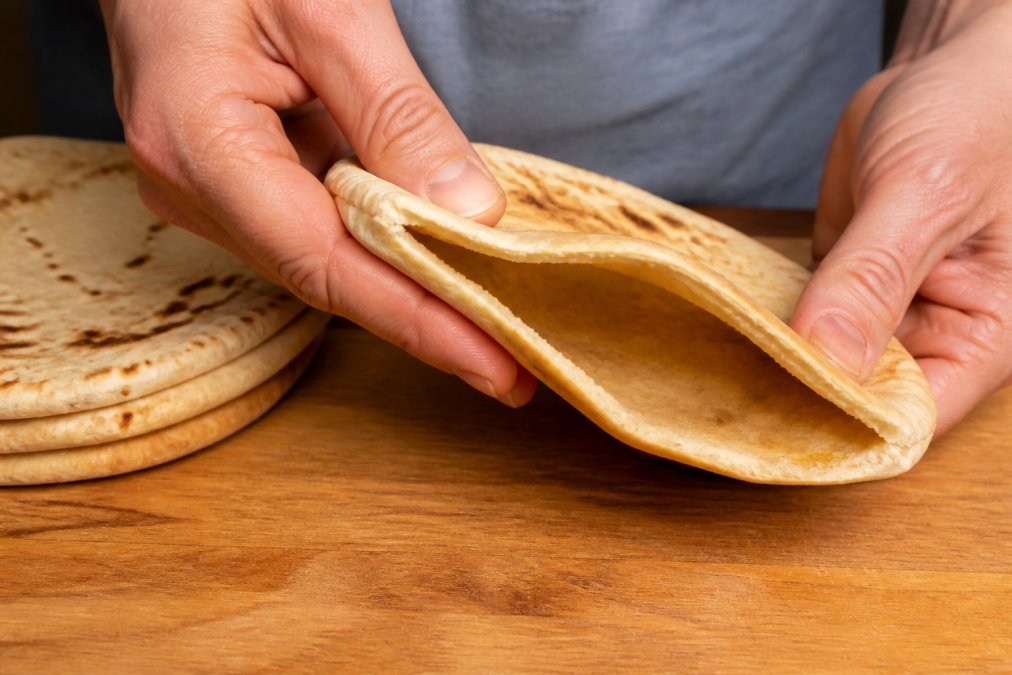 Close-up of hands gently pressing and folding a piece of fresh pita bread on a wooden kitchen counter, showing its soft, pliable texture and visible air pocket
