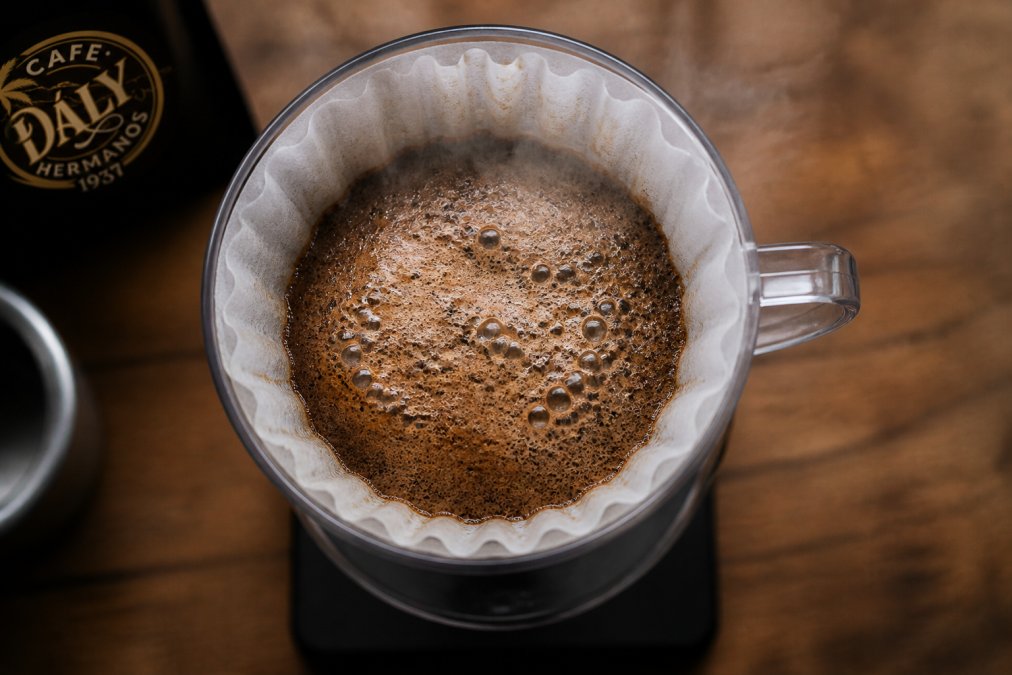 Overhead view of active coffee bloom in progress, showing thick layer of brown foam and visible bubbles rising from freshly ground coffee in a pour-over dripper setup with steam visible
