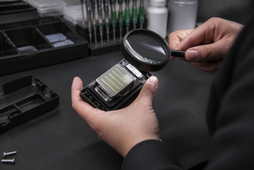 Close-up of a print engineer's hands holding a DTF printer printhead under bright workshop lighting, examining the nozzle plate and serial number label with a magnifying glass