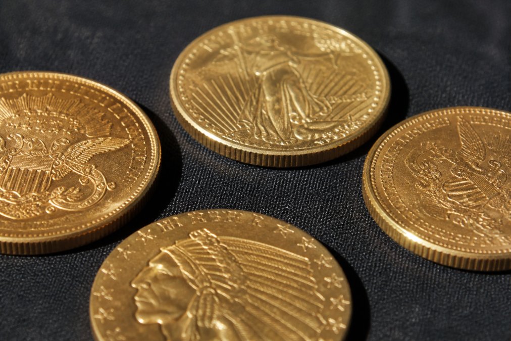 Close-up of several gold coins laid flat on a dark velvet surface, showing visible hallmarks, mint marks, and edge reeding details under bright directional natural light