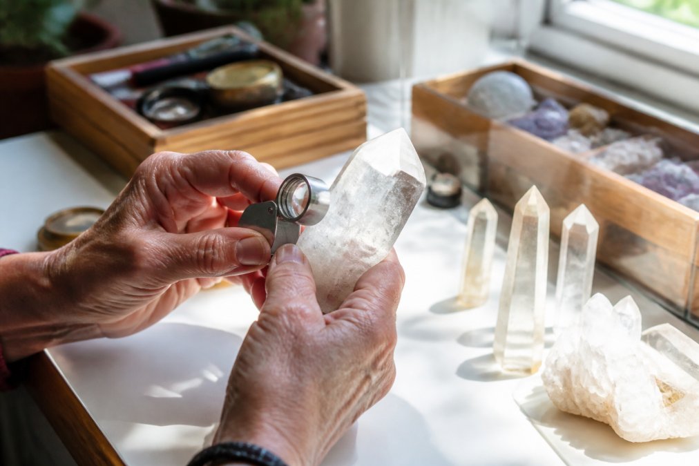 Jeweler's hands holding a magnifying loupe over a clear quartz crystal specimen on a white background, natural window lighting illuminating the stone from behind