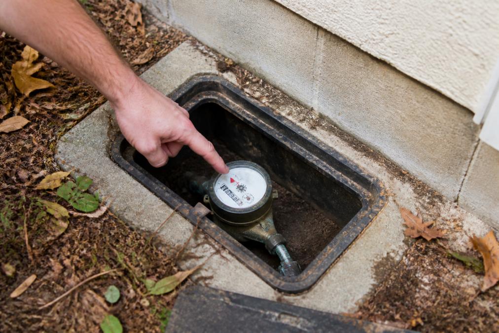 Homeowner's hand pointing to water meter dial with visible flow indicator in concrete meter box near home foundation, natural daylight