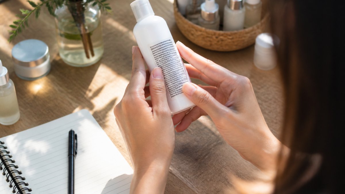 Close-up of hands holding a skincare product bottle, carefully examining the ingredient label text under bright natural light, with a small notebook and pen on the wooden surface nearby