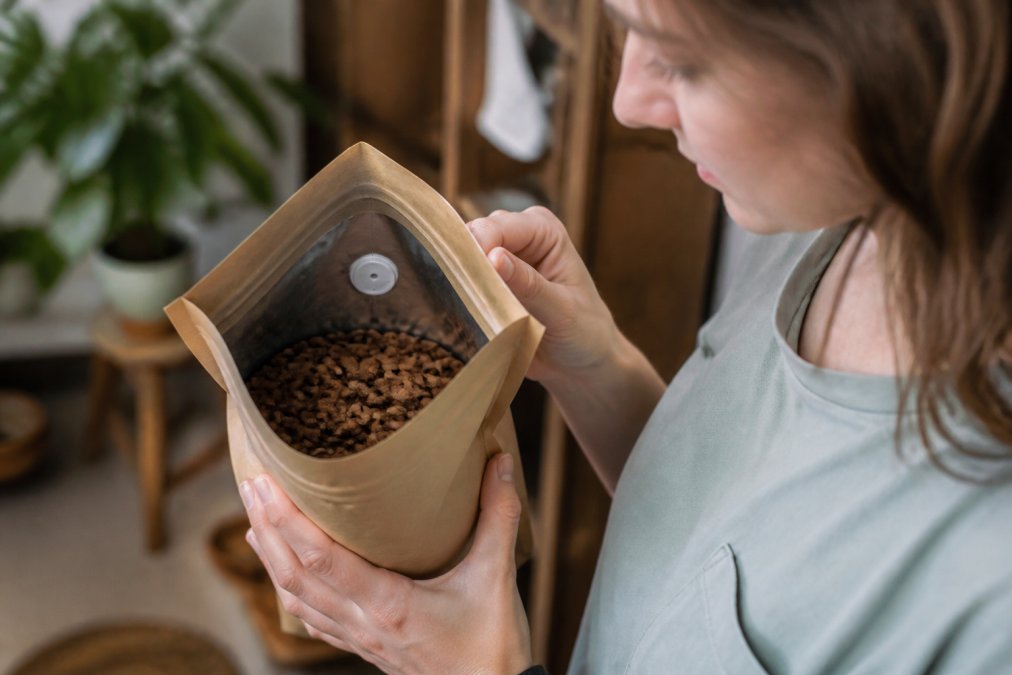 Hands holding an open kraft paper coffee bag with a visible silver one-way valve and a printed roast date label, revealing fresh medium-brown coffee beans inside