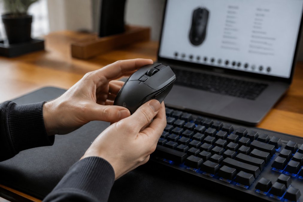 Close-up of hands holding a gaming mouse next to a mechanical keyboard on a desk, with a laptop showing a product spec sheet in the background