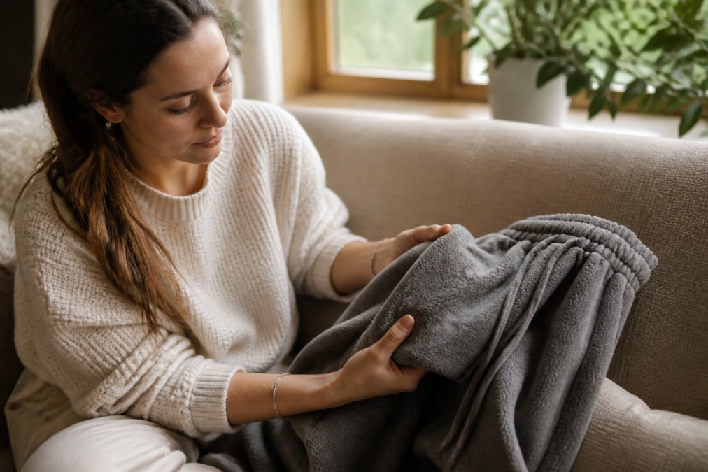 Close-up of a woman's hands gently pressing and feeling the soft fleece fabric of a pair of grey sweatpants, demonstrating the texture and density of the material in warm natural window light