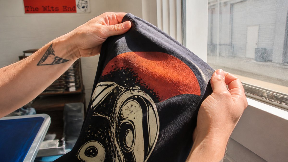 Close-up of hands holding a screen-printed T-shirt against natural light, examining the print edges and ink coverage in detail