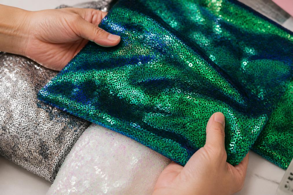 Close-up of hands holding different sequin fabric samples under bright studio lighting, showing texture and sheen variation between metallic and iridescent finishes on a neutral work surface