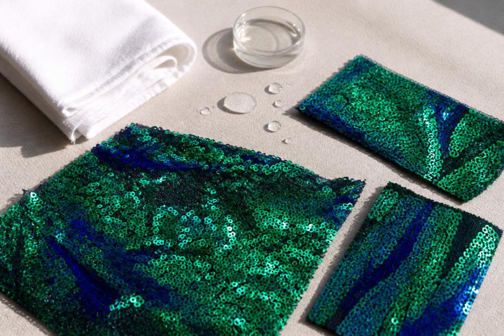 Flat lay of durability testing materials including white cotton rubbing cloth, water droplets on sequin samples, and iridescent sequin fabric swatches arranged on a neutral work surface with natural side lighting