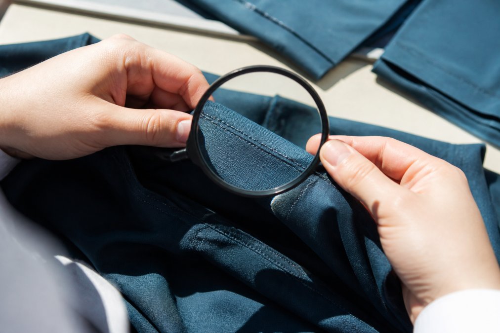 Close-up of professional hands examining a garment seam with a magnifying glass under bright natural light, showing straight stitching and thread alignment
