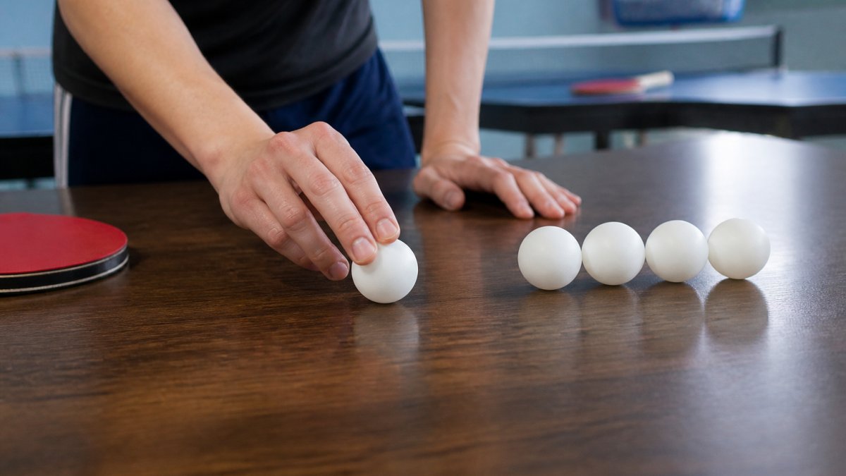 Close-up of hands rolling a white table tennis ball across a flat wooden table surface, with several other balls lined up nearby for comparison