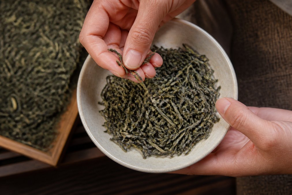Close-up overhead shot of whole leaf tea being examined in a small ceramic bowl, dry leaves held between fingers showing texture and uniformity