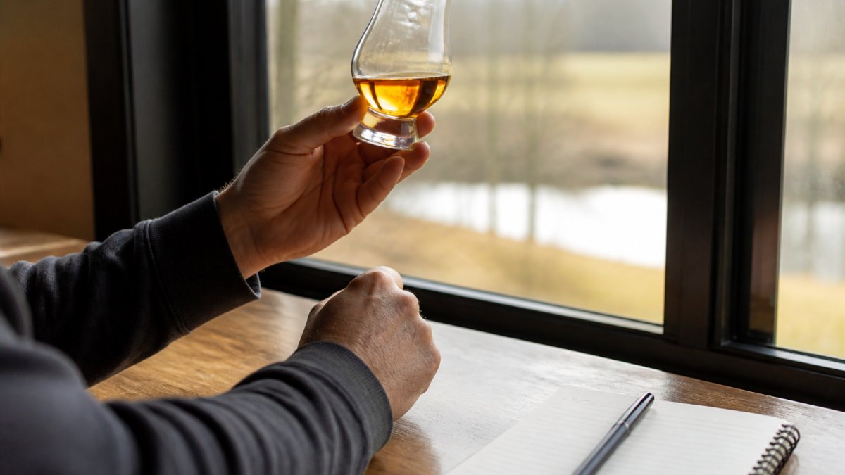 Close-up of a person's hands holding a Glencairn glass of amber whiskey up to a window, examining the color and legs, with a notebook and pen on the table beside them