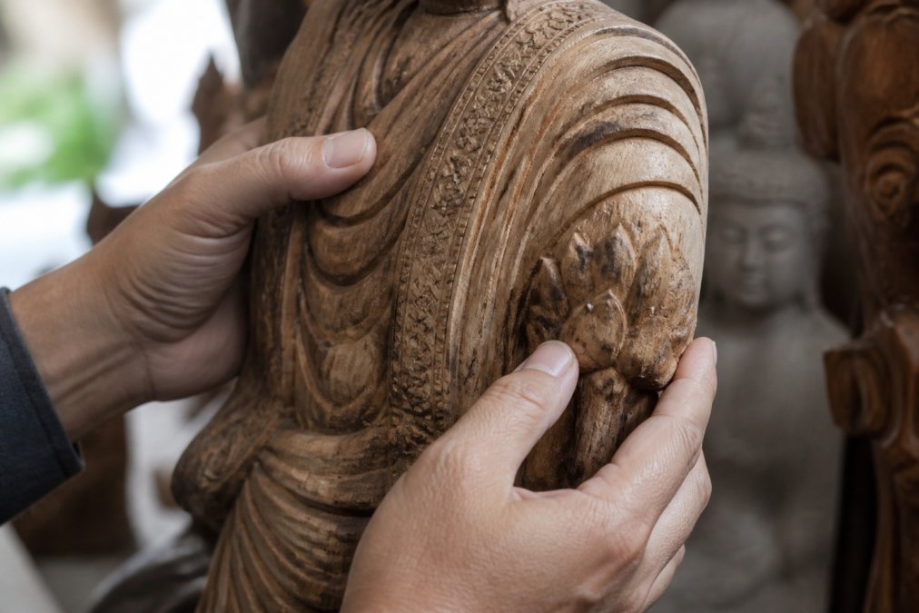 Hands carefully examining a wood carving detail, inspecting grain patterns and surface finish from multiple angles in natural daylight, showing close inspection of carved texture and wood characteristics