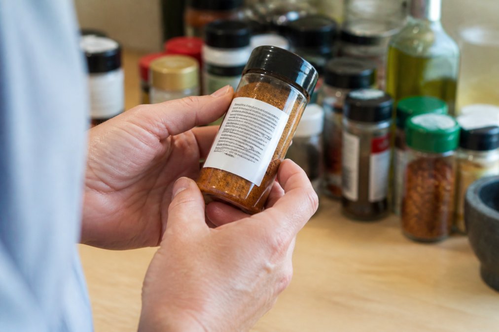 Close-up of hands holding a spice jar while reading the ingredient list on the back label, with natural kitchen lighting and other spice containers visible in soft focus in the background