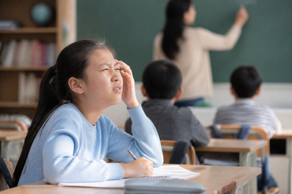 Child sitting at a school desk squinting at the classroom board while a teacher writes in the background, with natural classroom lighting highlighting the moment of visual difficulty