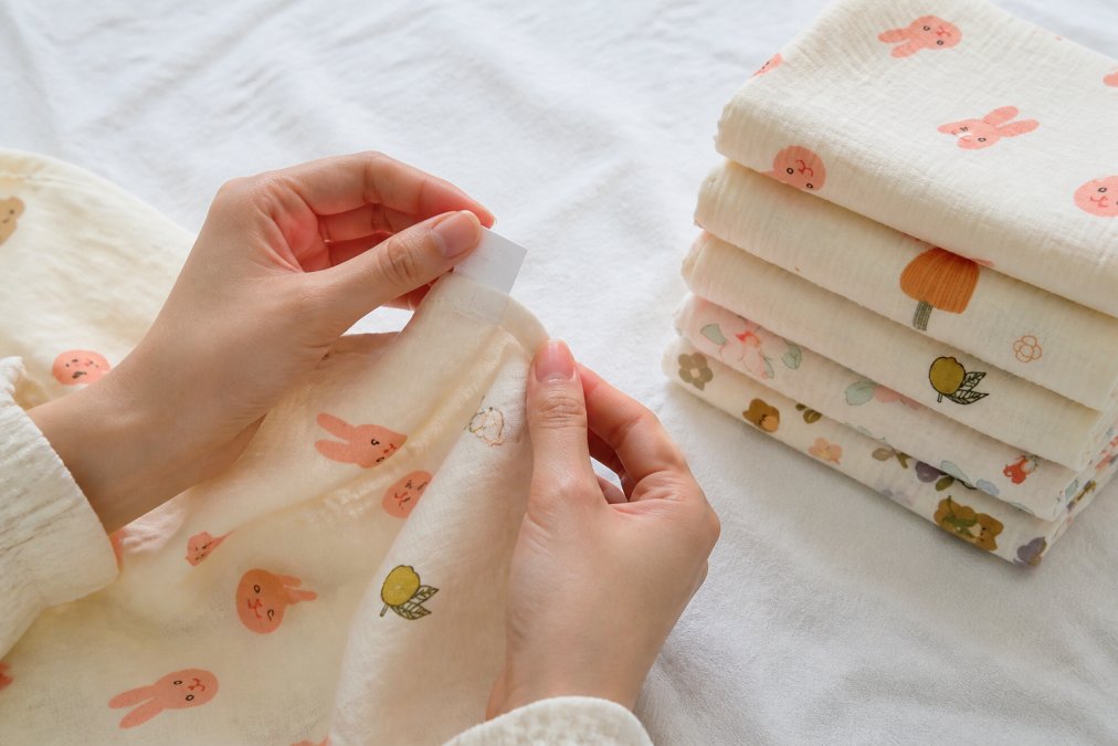 Close-up of a parent's hands holding a small baby garment and carefully examining the sewn-in care label tag, soft indoor light, white fabric background
