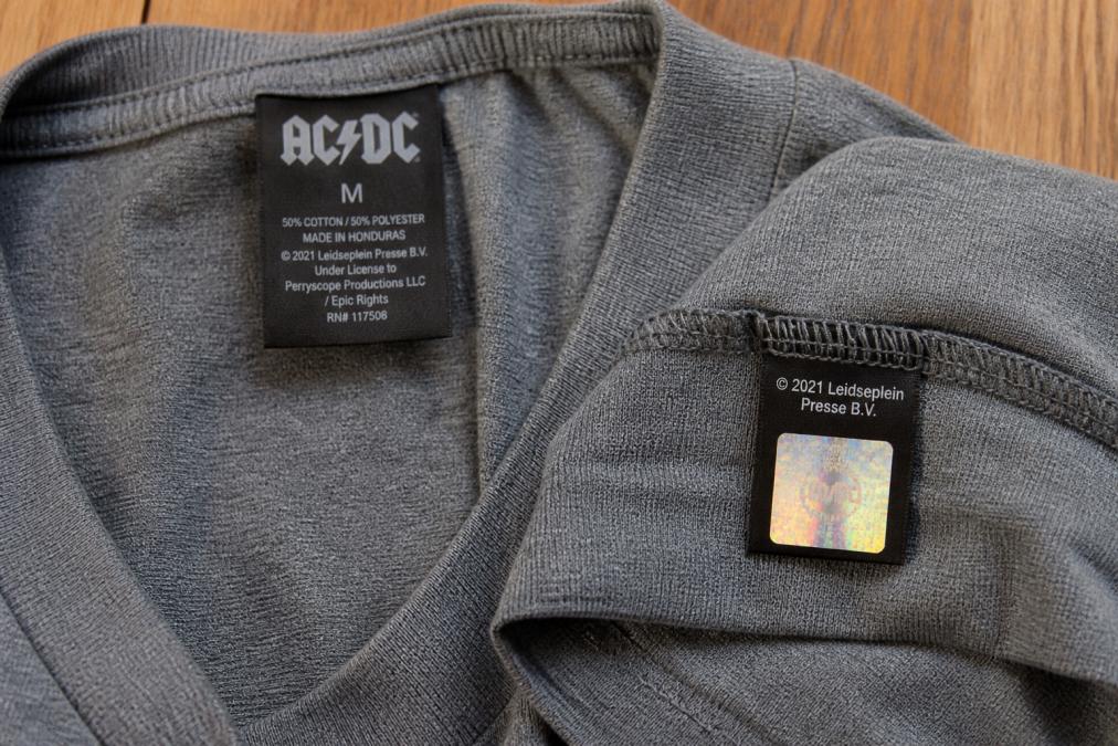 Close-up of the inside collar tag and hem label of a band t-shirt laid flat on a wooden surface under bright natural light, showing copyright text, a licensing label, and a small holographic sticker with visible color-shifting effect