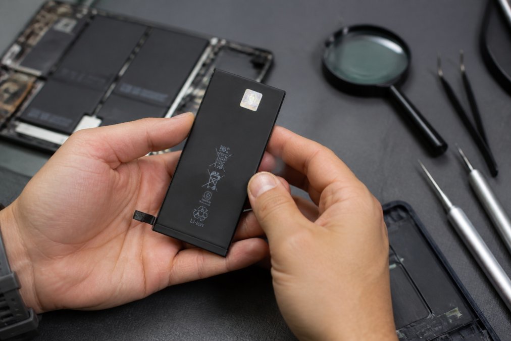 Close-up of a person's hands holding a smartphone battery with a visible OEM label and hologram, with a magnifying glass nearby on a clean workbench