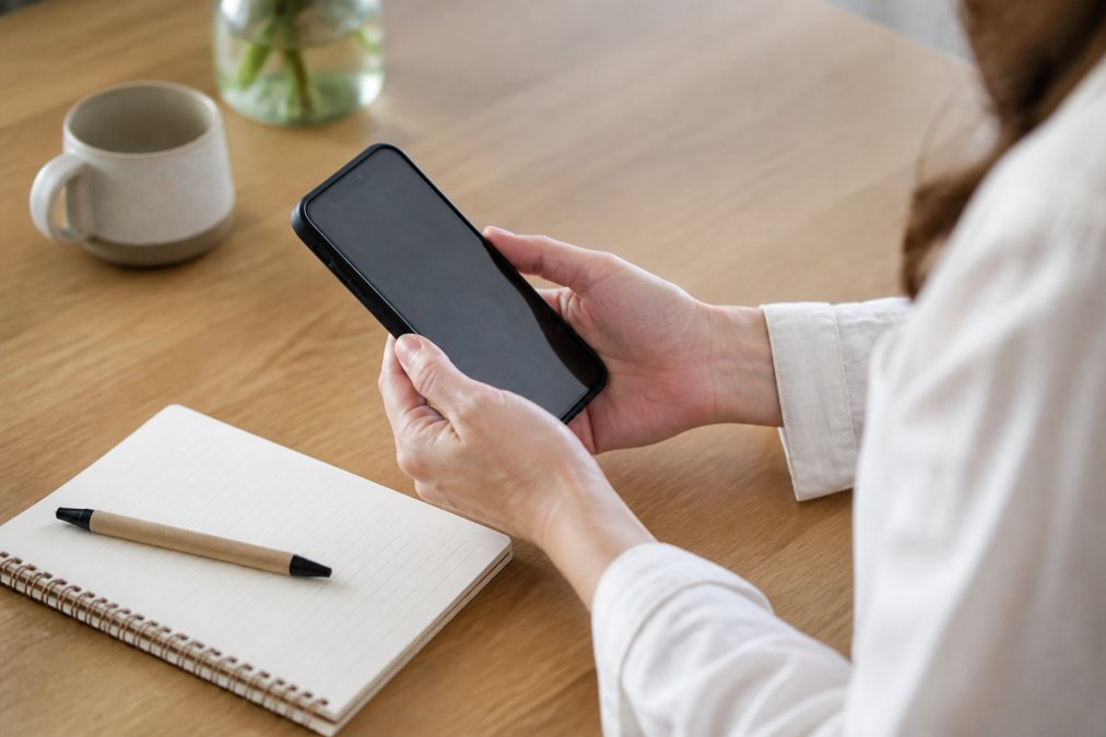 Close-up of woman's hands holding a smartphone displaying a health tracking app, with a notepad and pen nearby on a light wooden table