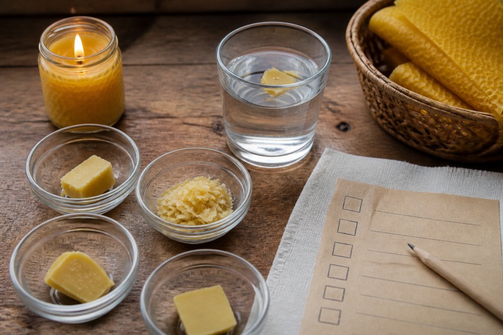 Close-up overhead shot of several small bowls of beeswax samples on a wooden kitchen counter, one piece floating in a glass of water, a lit candle nearby, and a handwritten testing checklist on paper beside them