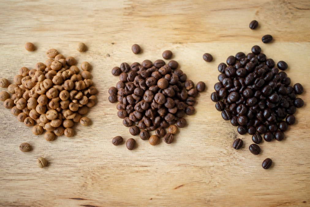 Close-up overhead shot of three small piles of whole coffee beans on a light wooden surface, showing visible color differences between light, medium, and dark roast levels, with a few beans scattered naturally around each pile