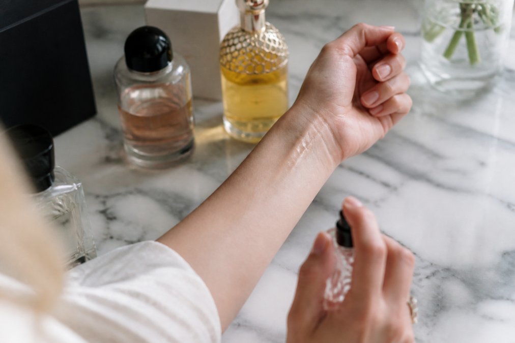 Close-up of a person's inner wrist with a small droplet of fragrance just applied, two elegant dark glass perfume bottles resting on a white marble surface in soft natural window light