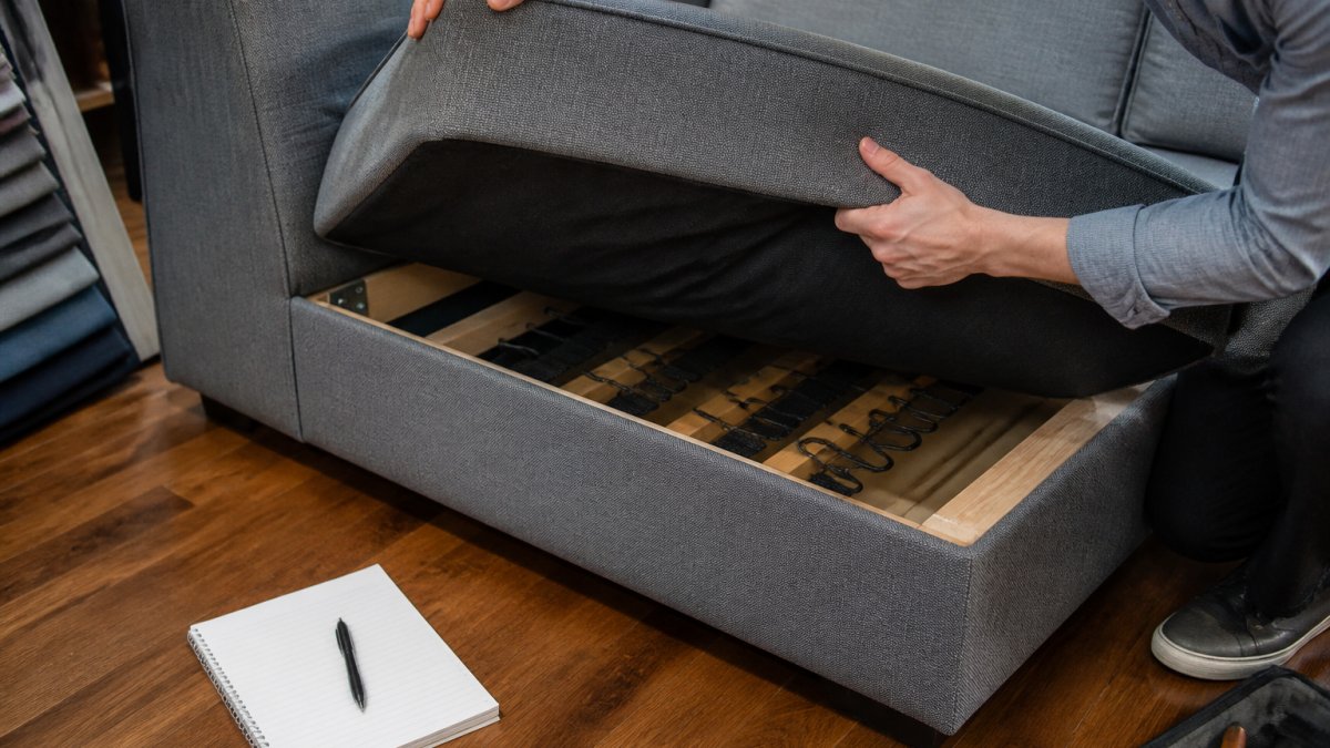 Close-up of hands lifting a sofa cushion to inspect the frame and suspension underneath, with a notebook and pen nearby on a hardwood floor