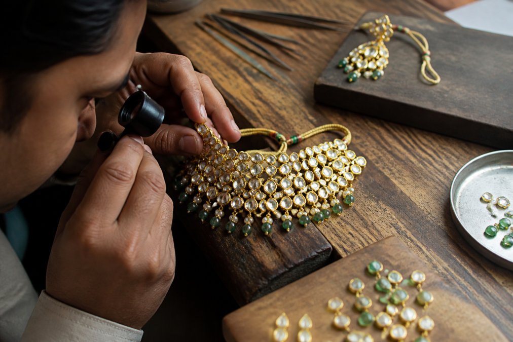 Close-up of hands examining a Kundan necklace under a jeweller's loupe at a wooden workbench, with soft daylight revealing uncut stones and gold foil setting