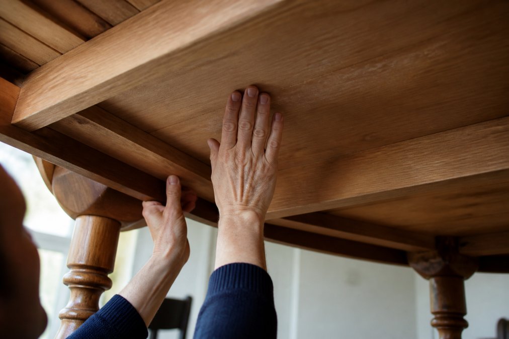 Close-up of a person's hands pressing and examining the underside of a wooden dining table, inspecting the raw unfinished wood grain in natural light