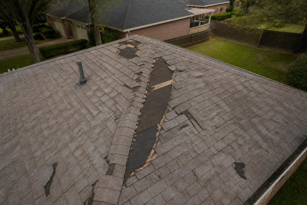 Overhead view of a residential suburban roof with several asphalt shingles completely missing near the ridge, lifted shingle edges exposing dark underlayment beneath, photographed in overcast natural light