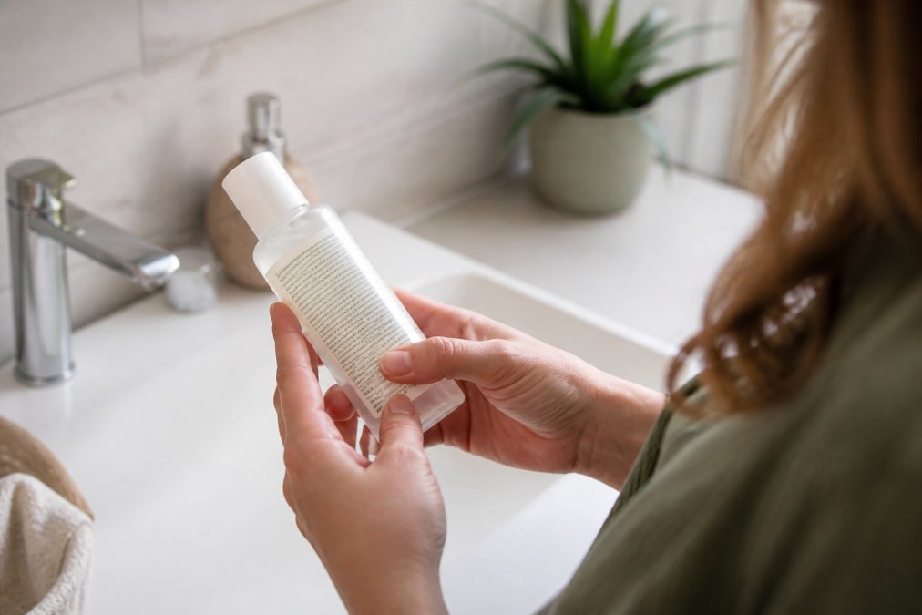 Close-up of a woman's hands holding a skincare bottle, turning it to read the ingredient list on the back label in a bright bathroom setting