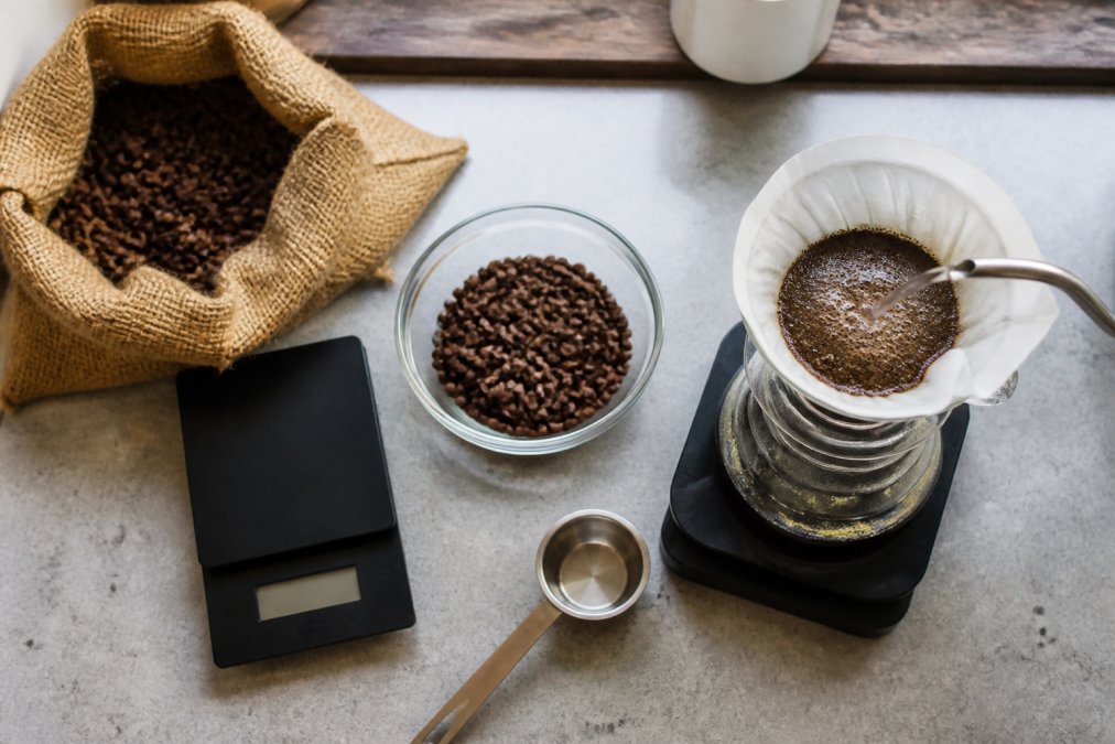 Overhead shot of a home kitchen counter with a digital scale, a burlap coffee bag, a glass bowl of whole roasted beans, and a hand pouring hot water over grounds in a pour-over dripper to show the bloom