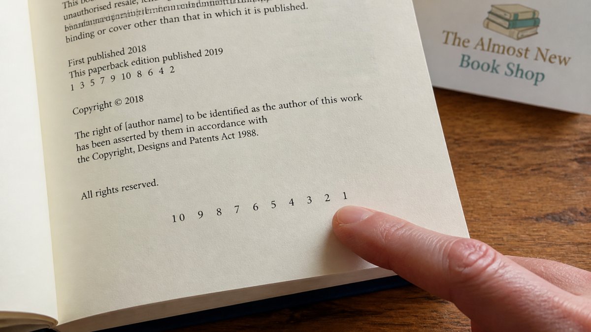 Close-up of an open book's copyright page showing the number line (sequence of numbers 10 9 8 7 6 5 4 3 2 1) with a finger pointing to the lowest number, illustrating how to read printing information