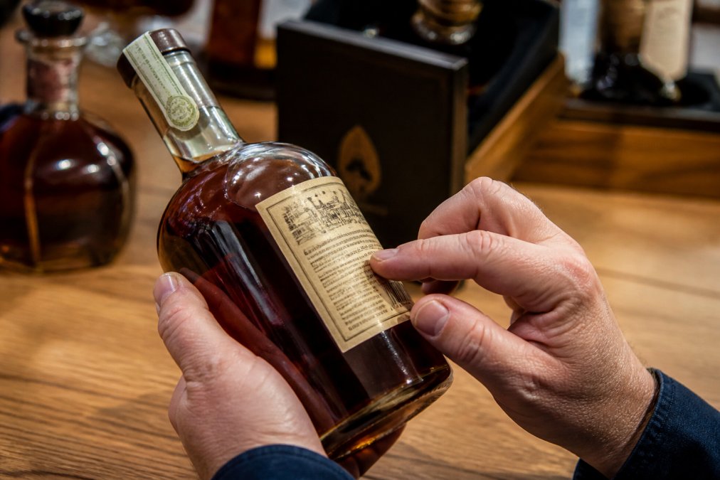 Close-up of hands examining a whiskey bottle label and seal under bright lighting, showing details of bottling information and authenticity markers