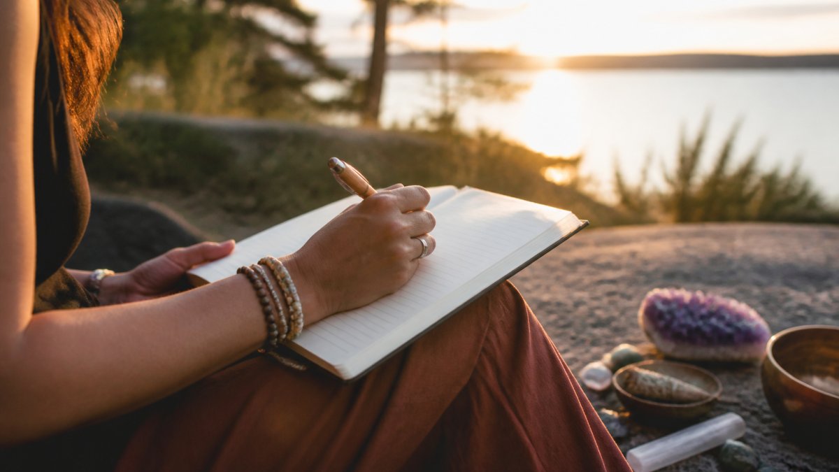 Person sitting peacefully on a forest path at sunset, journal in lap, pen in hand, gazing thoughtfully into the distance with golden light filtering through trees