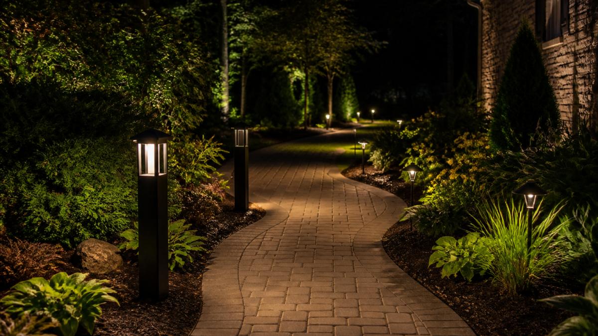 Residential garden pathway illuminated at night with evenly spaced pathway lights creating clear visibility along the walking surface, showing bollard lights and path lights positioned along garden beds and edges