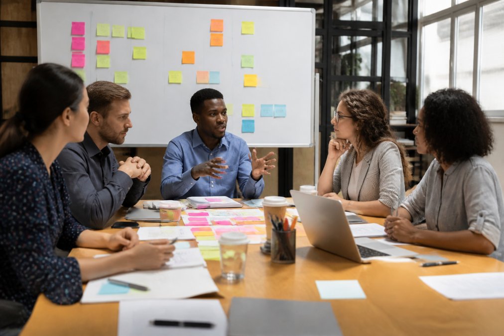Diverse team of professionals collaborating around a table with sticky notes, whiteboards, and laptops, discussing sprint planning in a bright, modern office environment