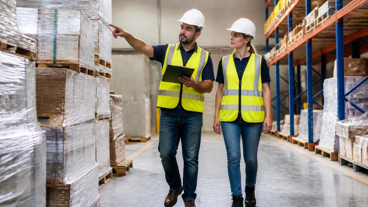 A small business owner and a safety consultant in hard hats walking a warehouse floor with a clipboard, pointing at a stacked pallet and checking a hazard checklist
