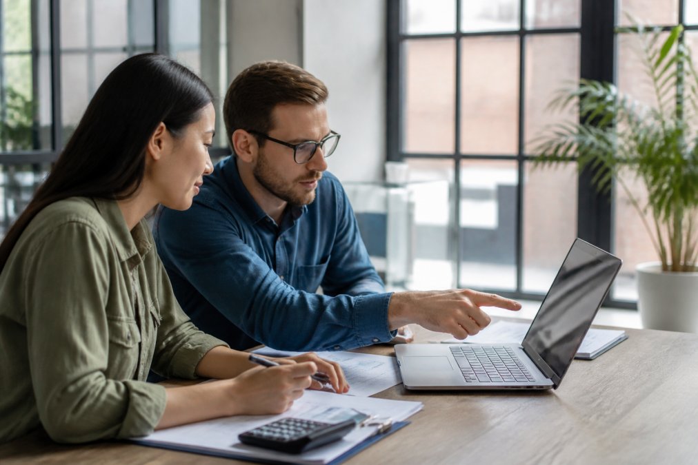 Two professionals reviewing financial reports and spreadsheets together at a table, pointing at a laptop screen showing budget data, bright modern office setting