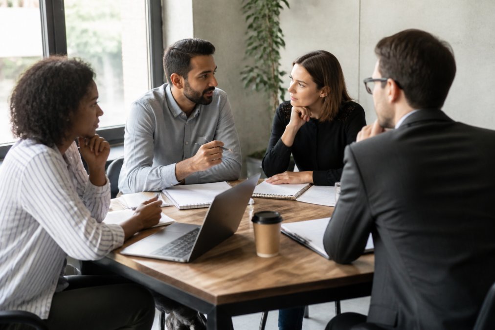 Small business team gathered around a table with notebooks and laptops, discussing strategy and action items in bright office setting with natural light