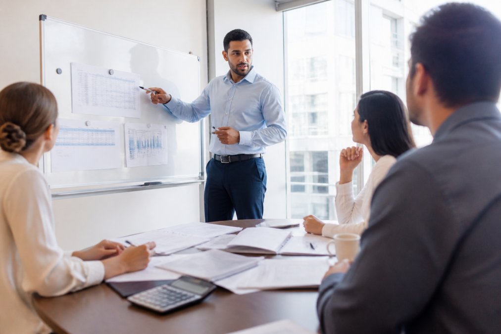 Business professional presenting pricing strategy to a team in a modern office conference room, pointing to strategy notes on a whiteboard, bright natural lighting