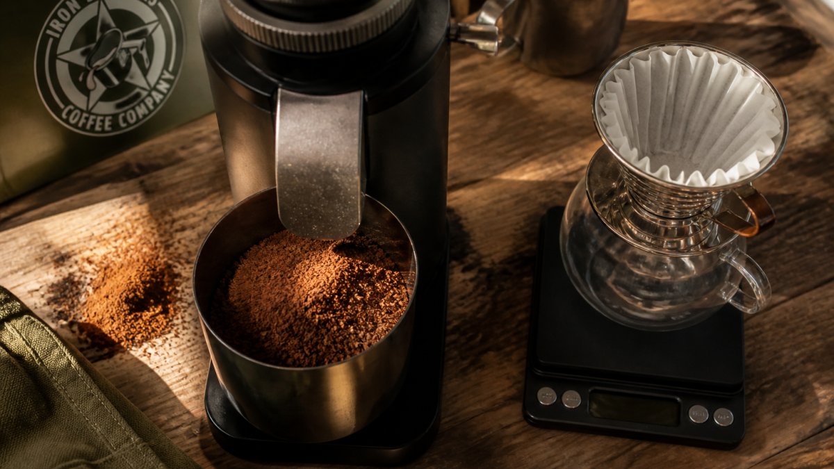 Overhead close-up of a burr grinder with freshly ground coffee in the catch cup, a small digital scale and a pour-over dripper on a wooden countertop, warm morning light