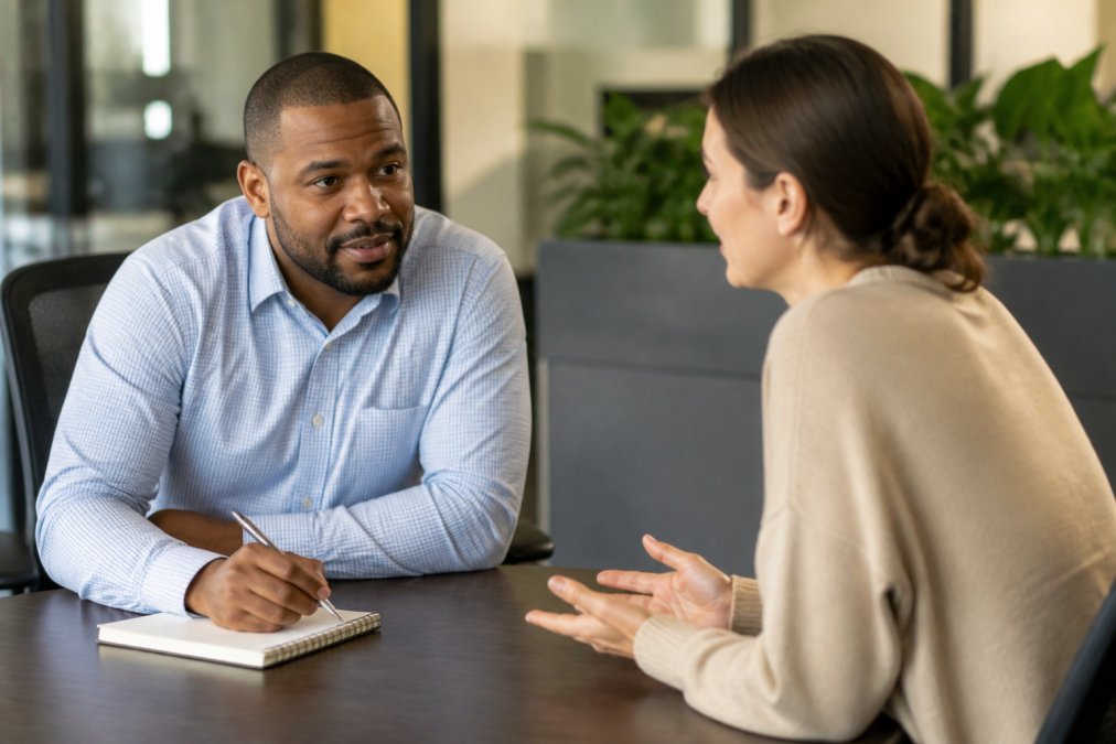 Manager sitting across from employee at desk, leaning forward with engaged posture, taking notes during one-on-one conversation in modern office setting