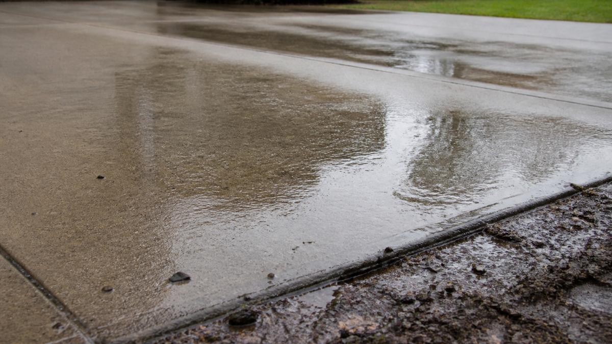 Close-up of standing water pooling on concrete driveway surface with muddy ground visible around edges during rainfall, showing water accumulation in low spot